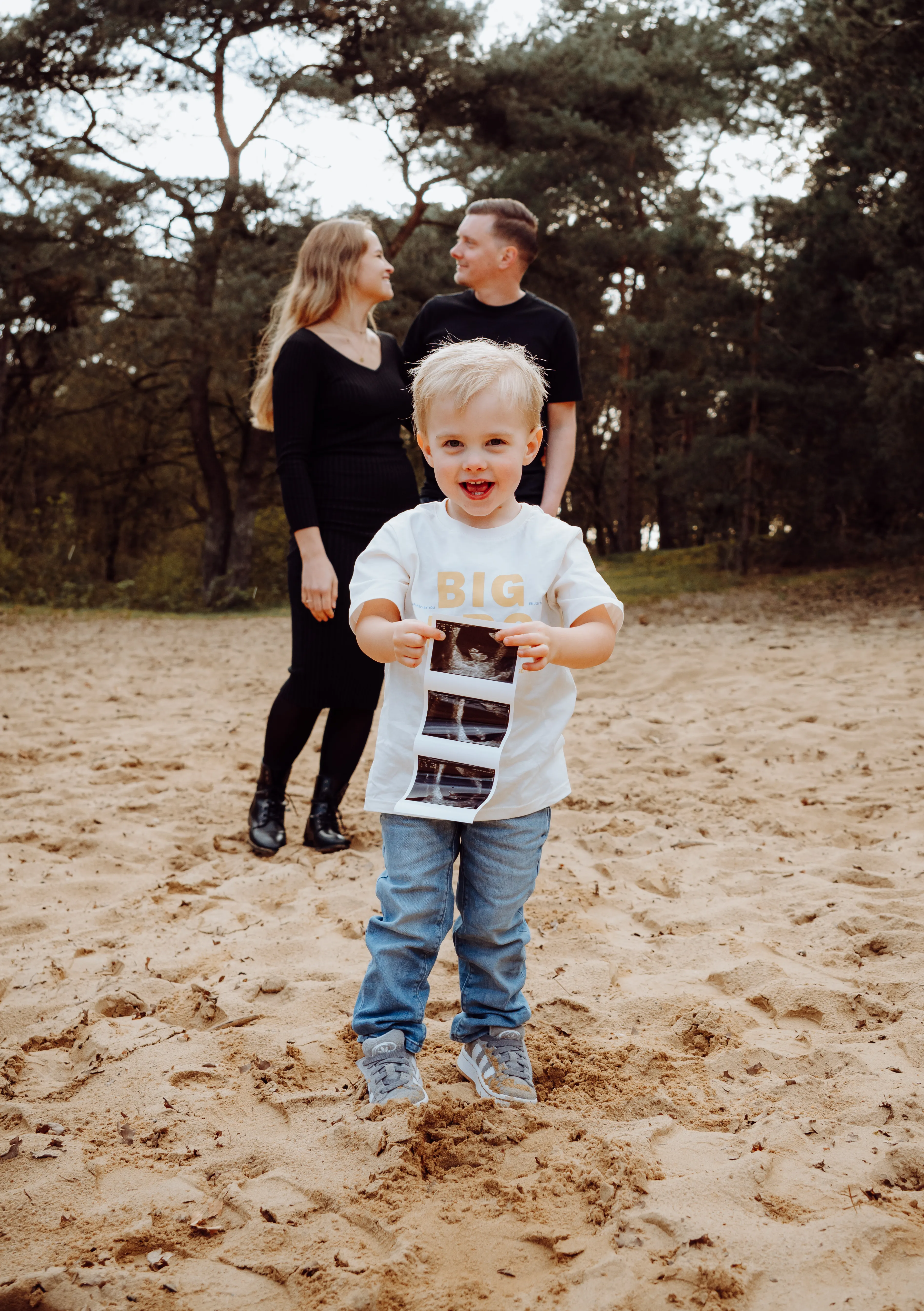 Zoontje houdt de echo omhoog terwijl zijn lachende ouders achter hem staan op een zandpad bij het bos