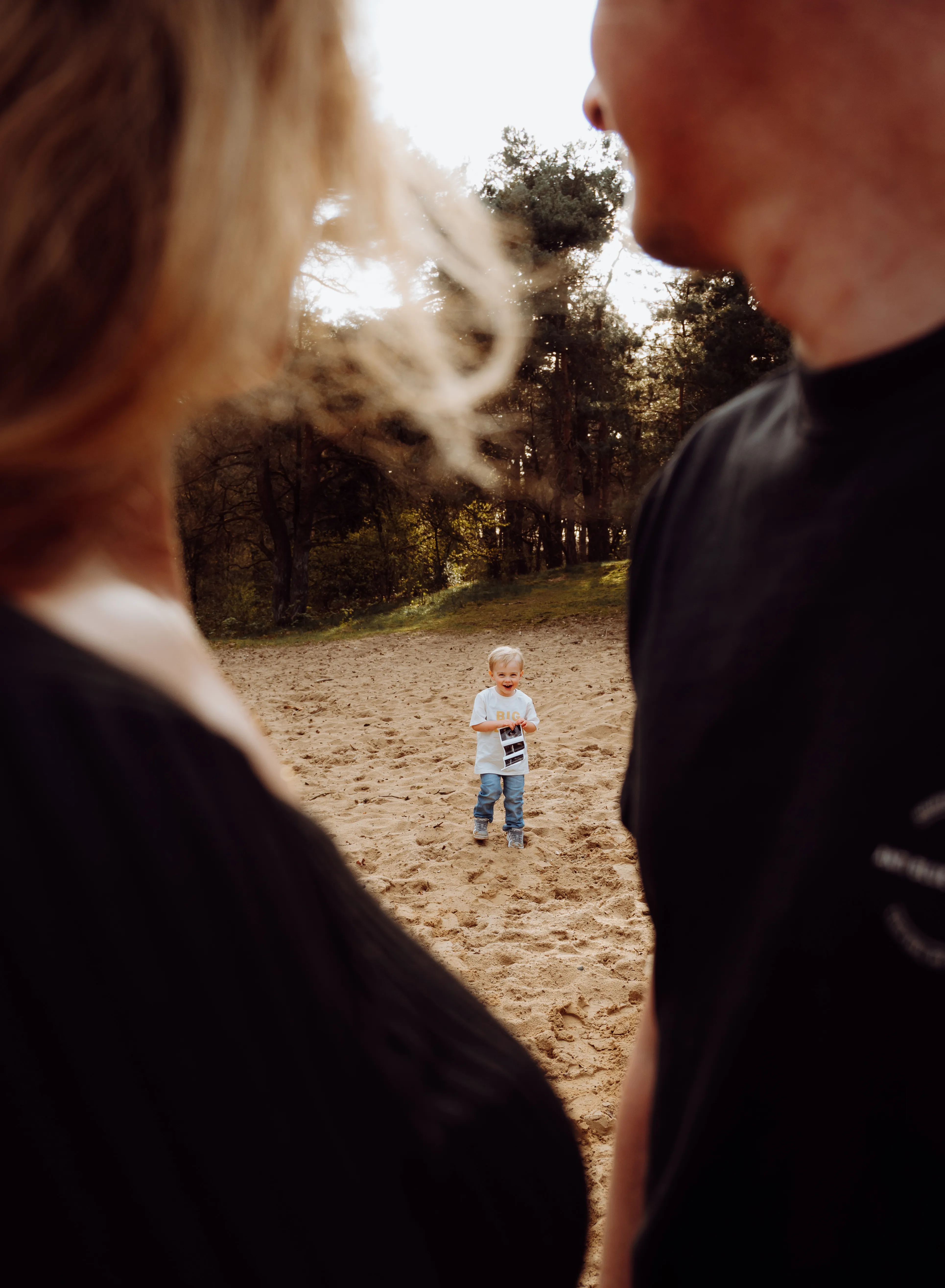 Lachende peuter op een zandpad tussen de dennen, omlijst door zijn vervaagde ouders in warm tegenlicht