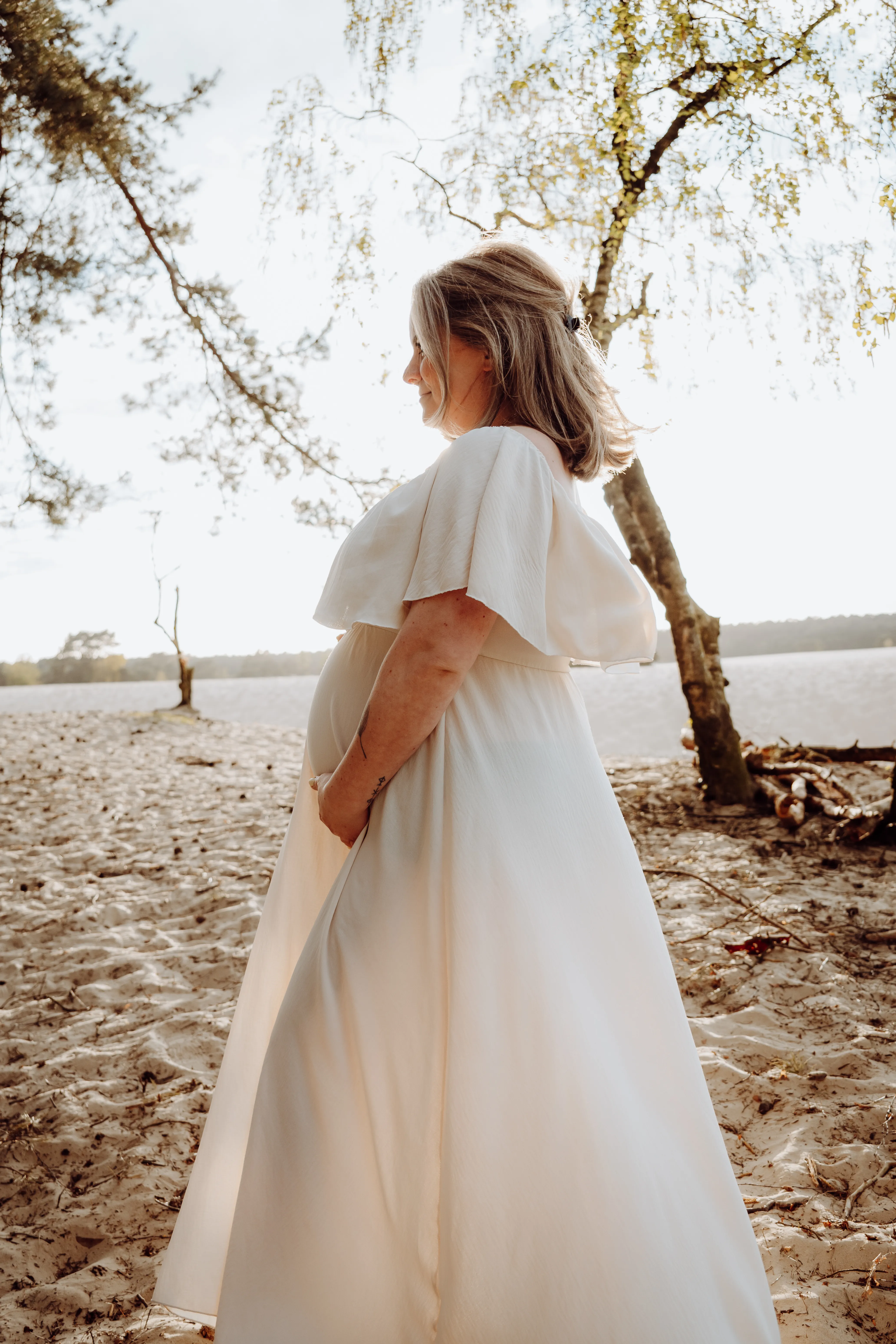 Zwangere vrouw in een lange witte jurk staat met haar hand op haar buik aan de waterkant in warm licht