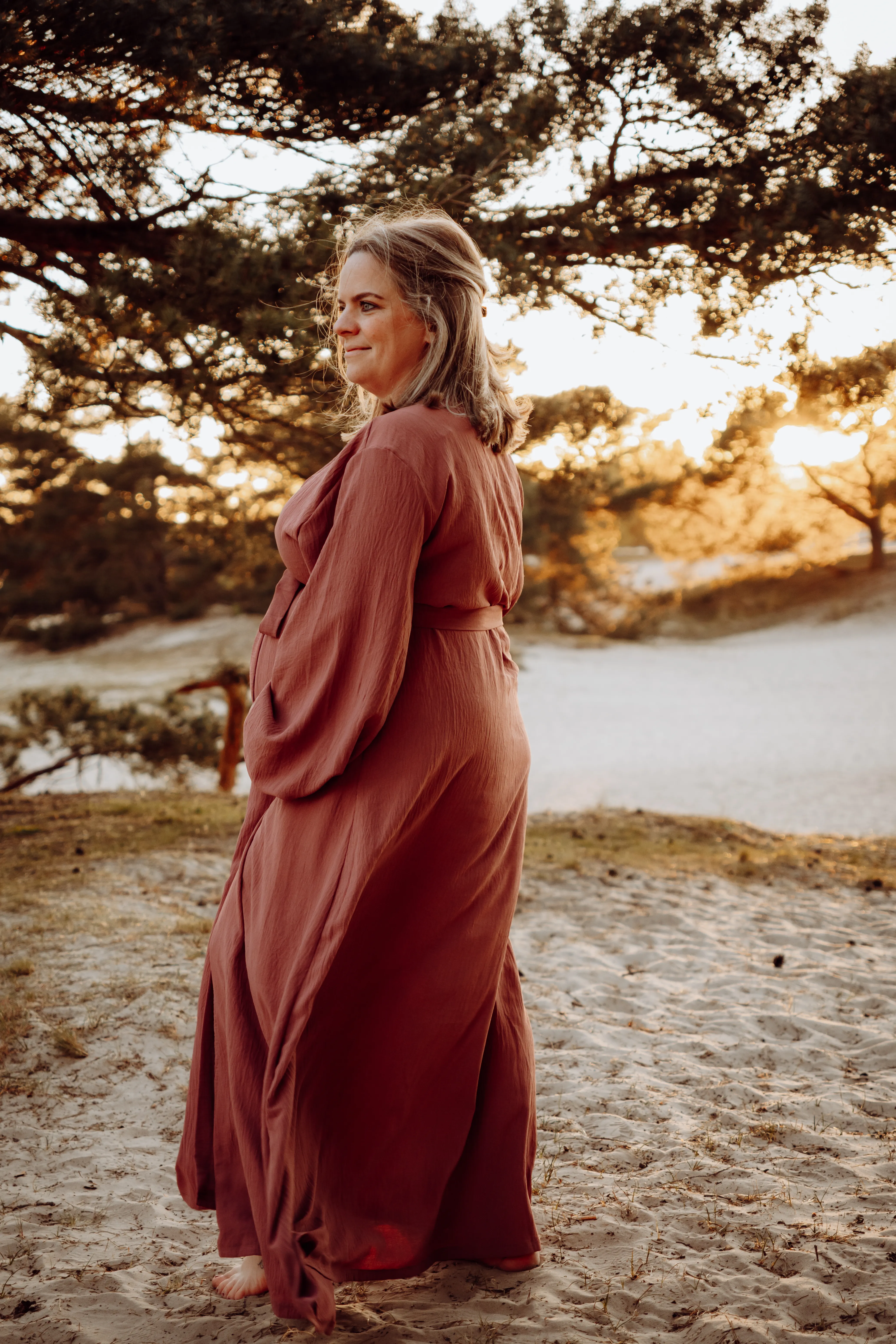 Zwangere vrouw in een lange terracotta jurk kijkt over haar schouder op het strand tijdens golden hour