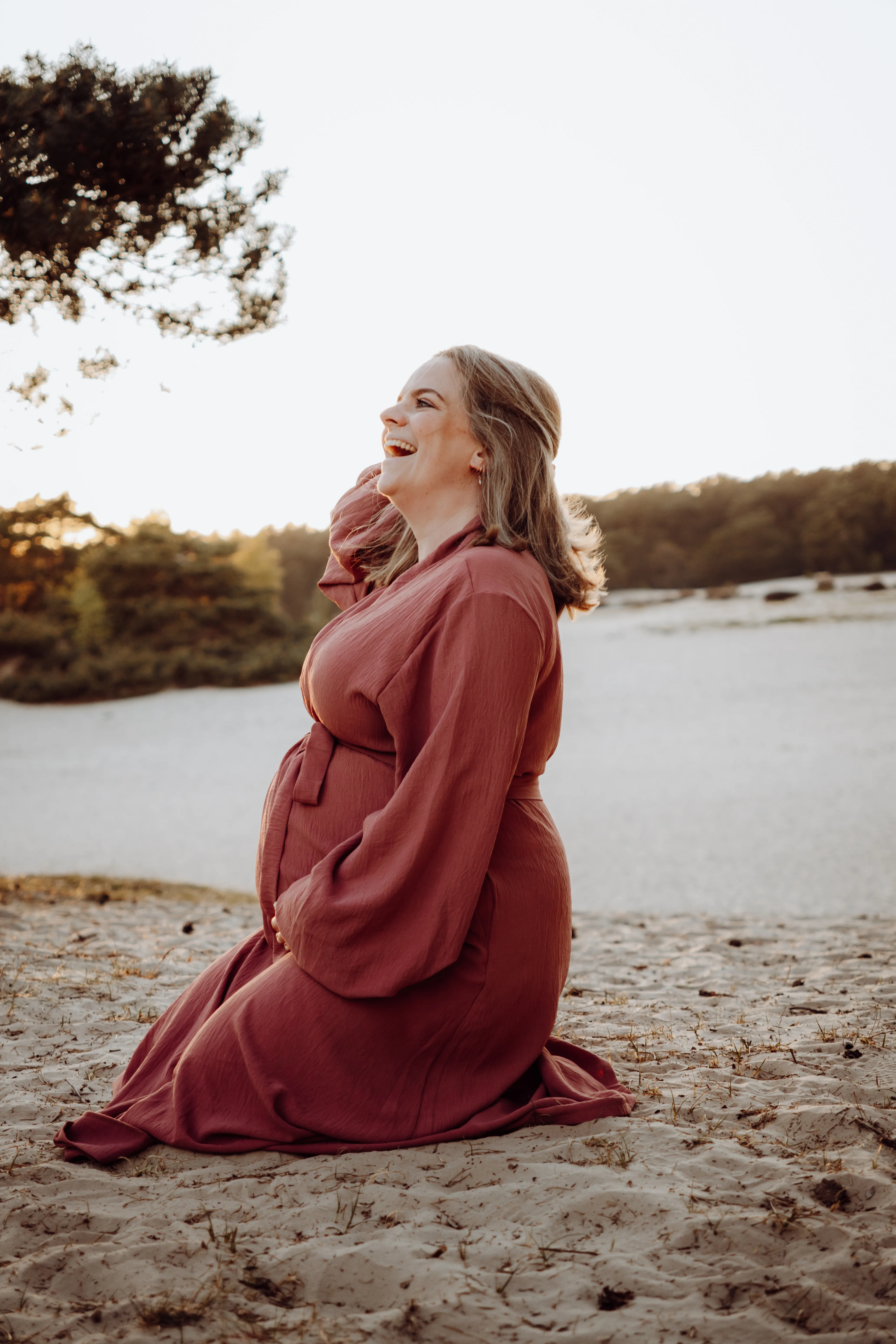 Lachende zwangere vrouw knielt in een terracotta jurk op het strand in warm avondlicht