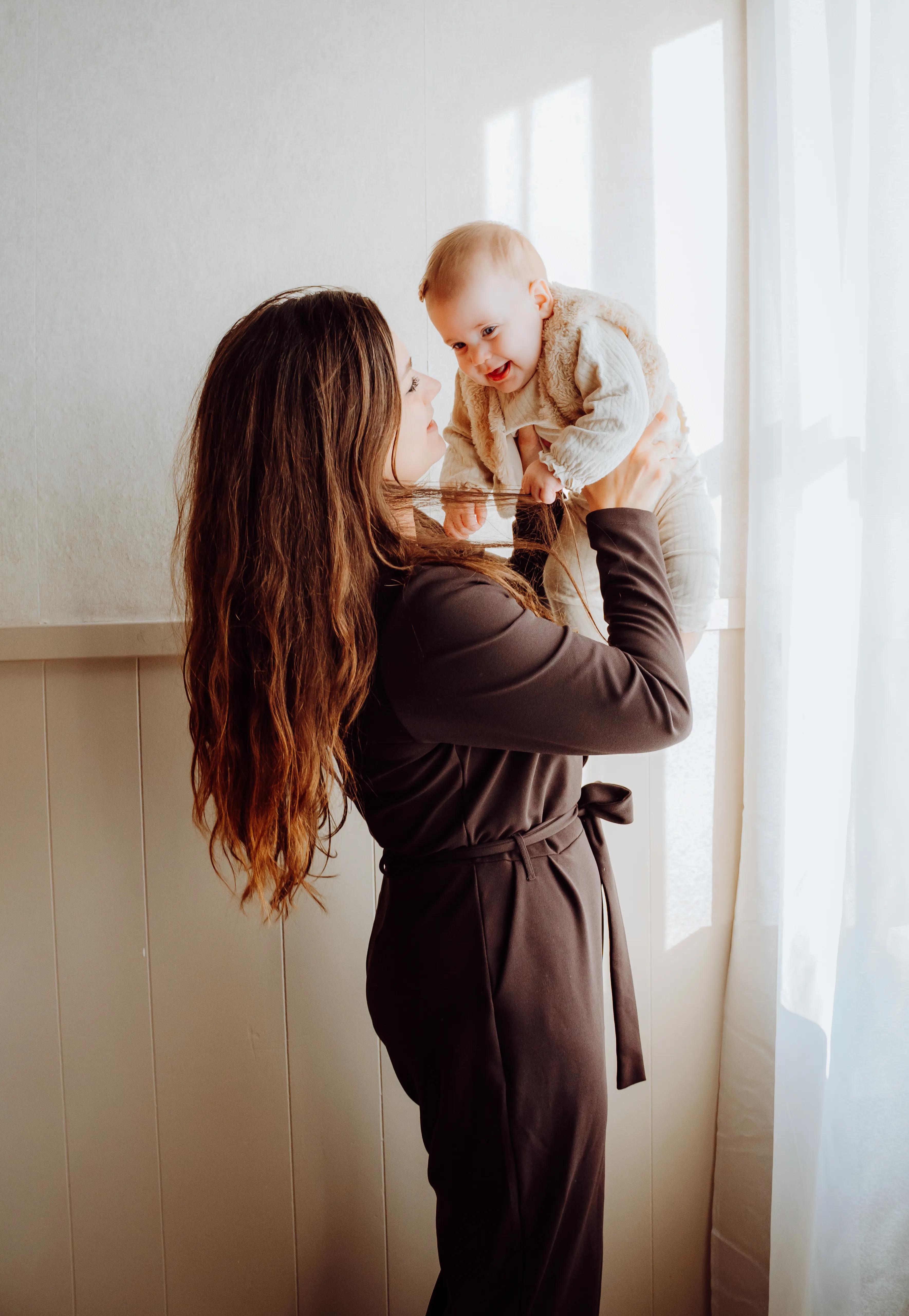 Vrouw tilt een lachende baby op bij het raam in zacht daglicht