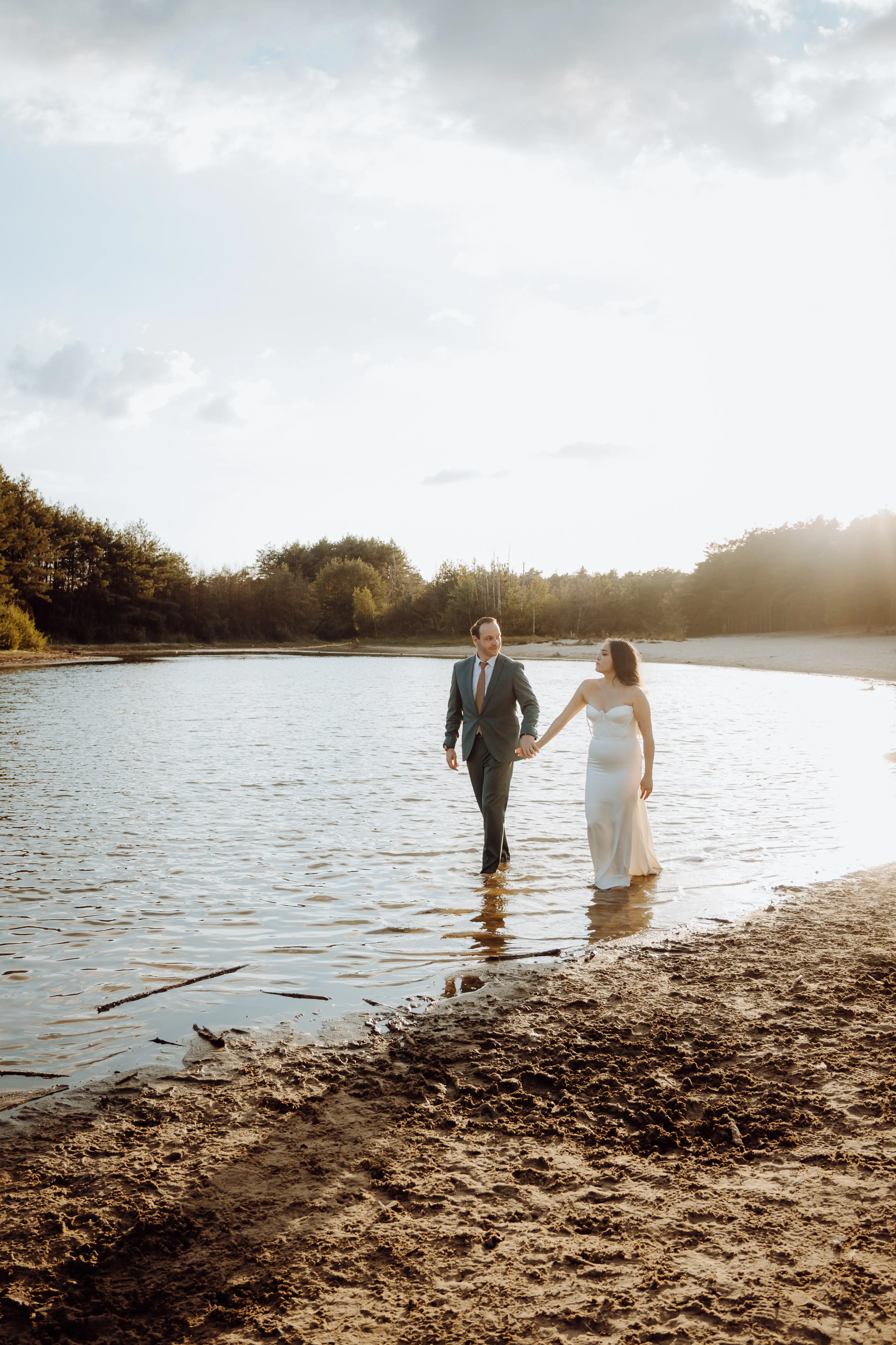 Stel loopt hand in hand het meer uit in warm gouden tegenlicht, met de modderige oever ervoor